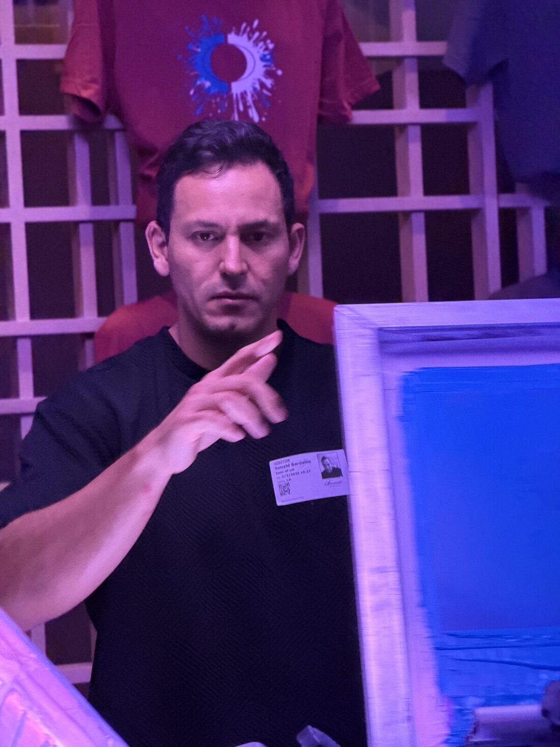 Crew member gesturing at a live station under violet event lighting with finished shirts displayed behind him